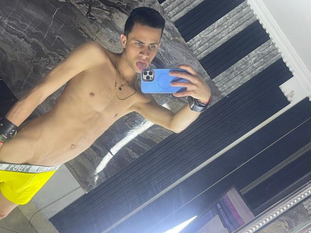 AxelBigCock, shirtless, yellow shorts, black wristbands, tongue out, holding blue phone, taking mirror selfie, short dark hair, necklace, slim build, reflection in mirror, standing in room.