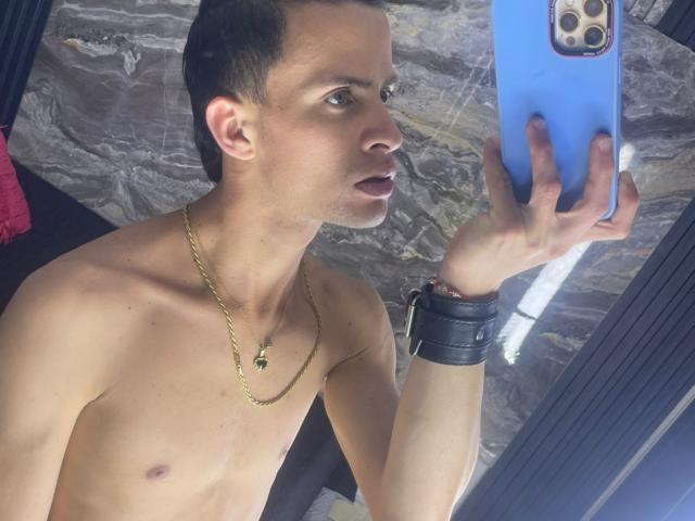 AxelBigCock, shirtless, with a gold chain, black wrist cuff, holding a blue phone, taking a mirror selfie, short dark hair, light skin, brown eyes, looking to the side.