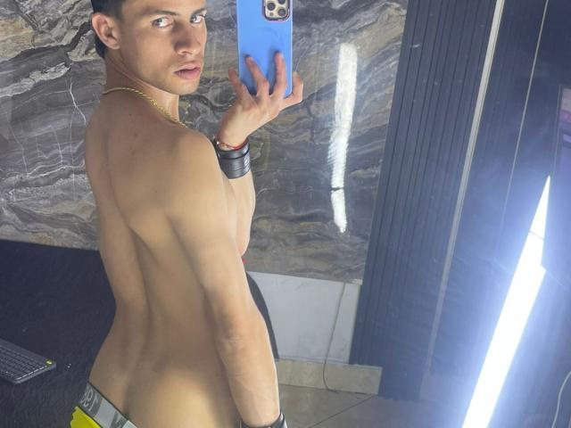 AxelBigCock, shirtless, muscular back, looking over shoulder, holding blue phone, black wristband, gold necklace, short dark hair, grey marble wall, mirror selfie, visible butt.
