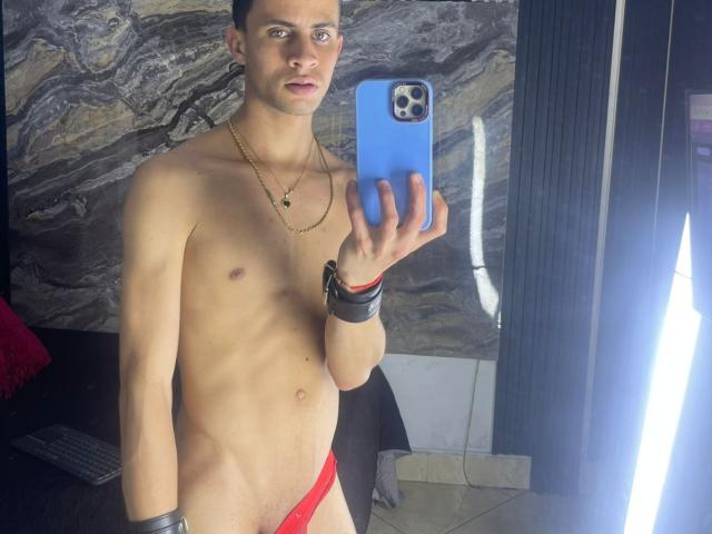 AxelBigCock, a slim, shirtless dude with a red thong, holding a blue phone, wearing a gold necklace and black wristbands, taking a mirror selfie.