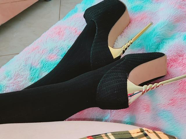 Akkanee's black textured high heels with gold stiletto heels are crossed on a pink, blue, and white fuzzy rug, showing off the shiny, pointed tips.