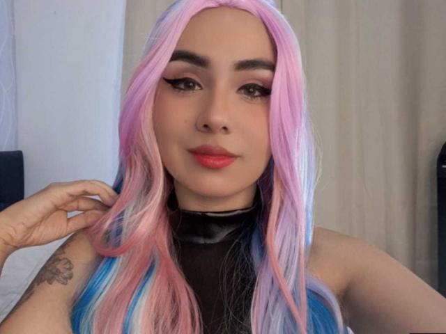 EmilyValk heeft roze en blauw haar, zwarte eyeliner, rode lipstick en een zwart topje zonder mouwen. Haar tatoeage zit op haar linkerarm. Ze kijkt naar de camera..