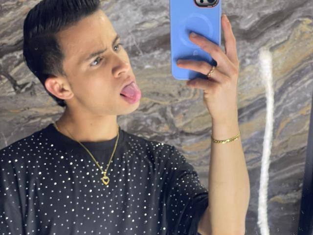 AxelBigCock with short black hair, tongue out, wearing a black sparkly shirt and gold necklace, holding a blue phone, wearing a gold bracelet, looking at the camera.
