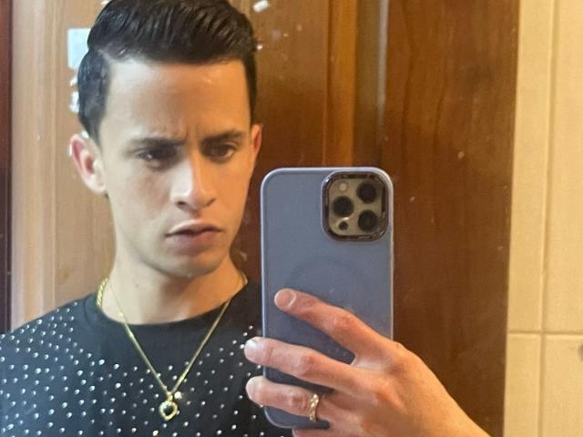 AxelBigCock with slicked-back dark hair, serious face, wearing a black polka dot shirt, gold necklace, holding a blue phone, taking a mirror selfie.