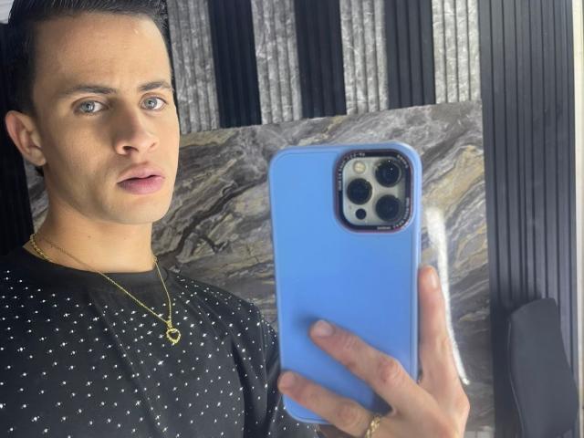 AxelBigCock with short dark hair, blue eyes, and a gold necklace, holding a blue phone with a marble pattern, wearing a black polka-dot shirt.