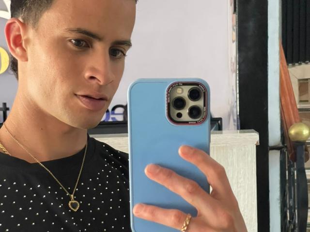 AxelBigCock with short dark hair, wearing a black polka dot shirt, gold necklace, and ring, holding a blue phone, taking a selfie, looking slightly to the side.