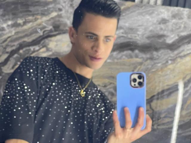 AxelBigCock with short black hair, blue eyes, wearing a black sparkly shirt, holding a blue phone, taking a mirror selfie, gold necklace, looking slightly to the side.