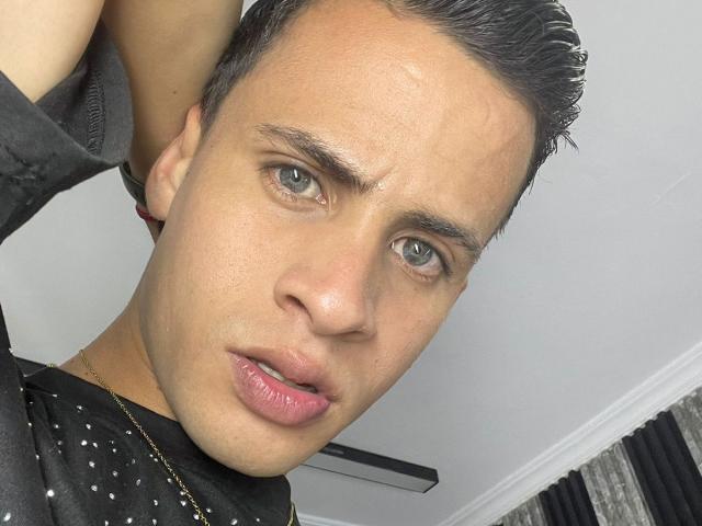 AxelBigCock with shiny green eyes, short dark hair, and a slightly parted pink-lipmed mouth, wearing a black shirt with small white dots. Hands behind head, looking at camera.