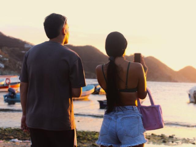 ZairaAndres stands with her back to the viewer, holding a purple bag in her right hand and a smartphone in her left. She has long dark hair in a ponytail and wears a black bra and blue denim shorts. A man with short dark hair, wearing a dark gray t-shirt and black pants, stands to her left. The sun sets over a calm, rocky shoreline with small boats in the water. Warm, golden light illuminates the scene.