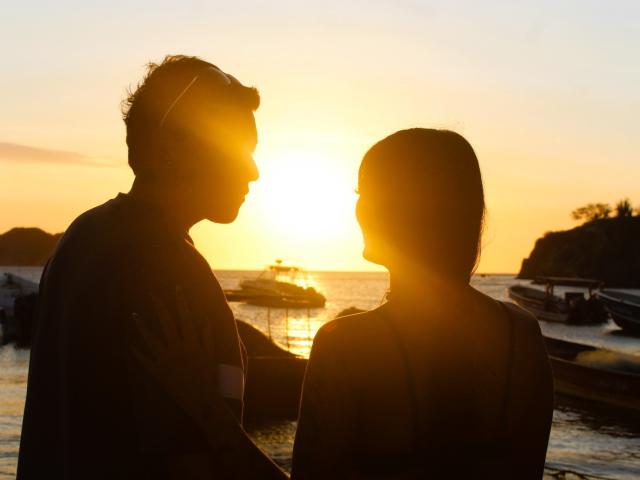 Silhouetted ZairaAndres with short hair and another ZairaAndres with curly hair, standing close, facing each other, sunset in background, hands touching, golden sun, calm water.