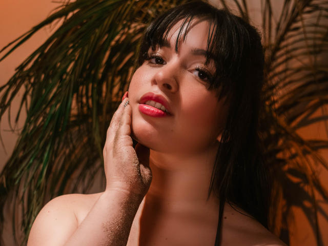KateLeeh with dark hair, pink lipstick, and a hand on her cheek, looking up with a slight smile, has a smooth, fair-skinned face and a small mole on her chin.