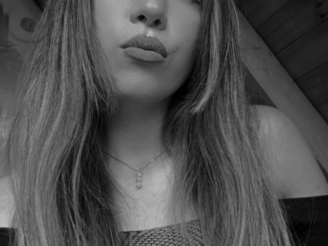 Black-and-white selfie of IzzysMisss with pouty lips, long straight hair, necklace, and off-shoulder top, showing her tits slightly. Her ass is not visible.