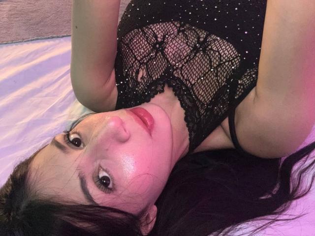SophieBeaumont with shiny skin, black lace top, and red lipstick, lying on a bed, head tilted, long dark hair, looking at the camera, pinkish light on her face.
