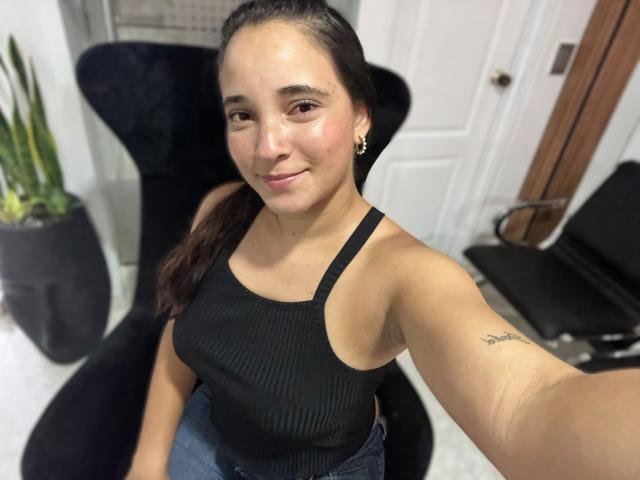 SophieMorgan with dark hair in a ponytail, wearing a black ribbed tank top, showing her tits and a tattoo on her right arm, smiling at the camera.