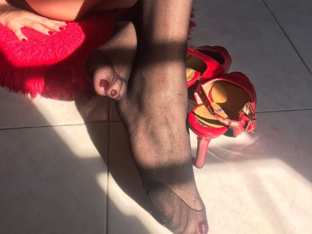 Close-up of VittoriaI's legs in black sheer stockings with red toenail polish, crossed on a white tiled floor. Red high-heeled sandals with a floral bow are to the right. She's sitting on a bright red, fluffy textured surface. Sunlight casts shadows across the legs and floor. The composition emphasizes the contrast between the dark stockings and the bright red elements.