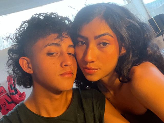 KeithAndBellaC with curly dark hair and green shirt, and KeithAndBellaC with wavy dark hair, nose ring, and no shirt, looking at camera, both with relaxed, slightly sleepy expressions.