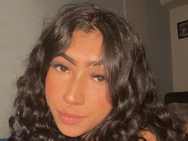 KeithAndBellaC has curly black hair, brown eyes, and a nose ring. Her glossy lips are slightly parted in a subtle smile. She's looking directly at the camera.