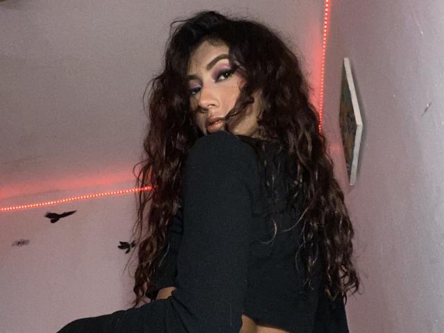 KeithAndBellaC with long curly brown hair, black long-sleeve top, looking back with bold makeup, red LED strip lights in background, serious expression, dark eyebrows, medium skin tone.