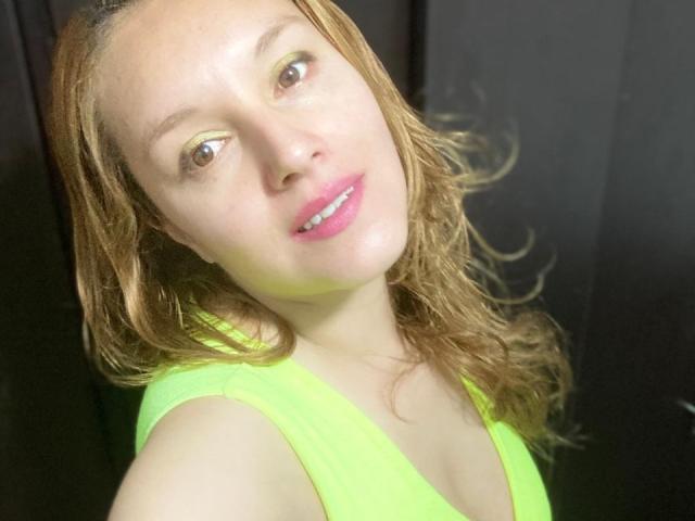 ChloeJewel with light brown wavy hair, green eyeliner, pink lipstick, and a neon green tank top, showing her tits and a slight smile in a selfie.
