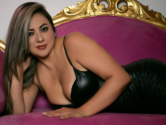 AmandaLeon with big boobs in a black leather dress, lying on a purple velvet couch, gold ornate headboard, looking at the camera with a slight smile.