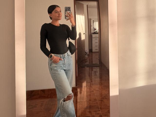 Thalia Ares, con cabello oscuro, una camiseta negra de manga larga y jeans azules rotos, está de pie frente a un espejo, sosteniendo un teléfono. El suelo es de madera y la habitación está iluminada por el sol. Su trasero es lindo..