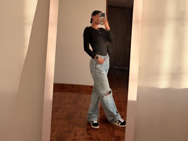 ThaliaAres in a black long-sleeve top, ripped light blue jeans, black and white sneakers, holding a phone, mirror selfie, wooden floor, sun shadows, casual, confident.
