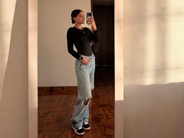 ThaliaAres with dark hair in a bun, wearing a black long-sleeve top, light blue ripped jeans, and black-and-white sneakers, standing in front of a mirror, holding a phone.