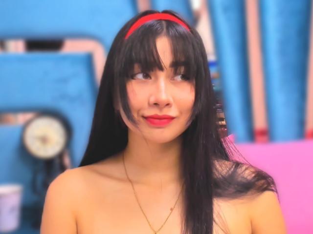 Genesiss has long black hair, red headband, red lipstick, and a gold necklace, showing a cute smile with her bare shoulders visible. Her boobs are slightly visible.