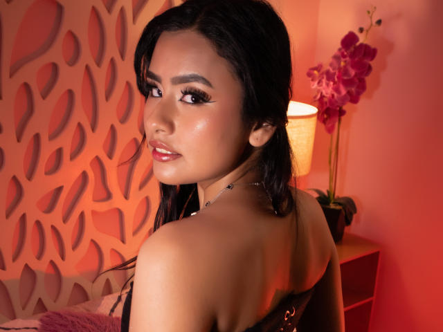 NelttDoll with glossy brown eyes, black hair, and shiny skin, looking back with a slight smile, showing her bare shoulders and black strap, next to a pink orchid.