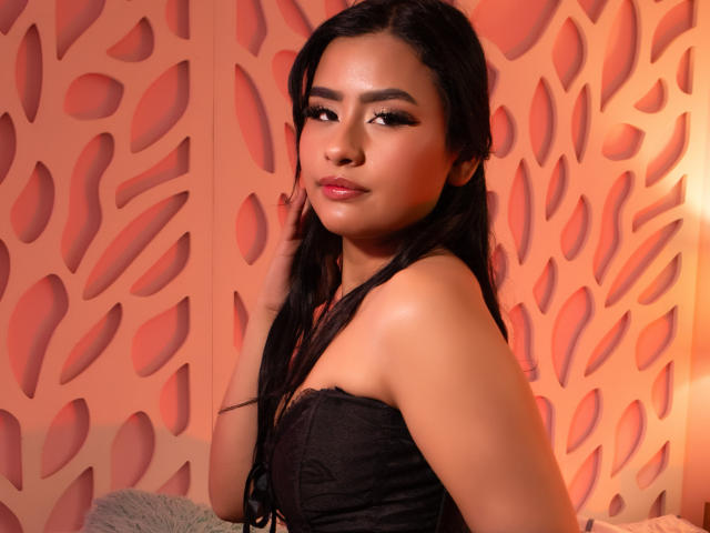 NelttDoll with long black hair, brown skin, and dark eyes, wearing a black lace top, has her hand on her cheek, looking over her shoulder with a slight smile.