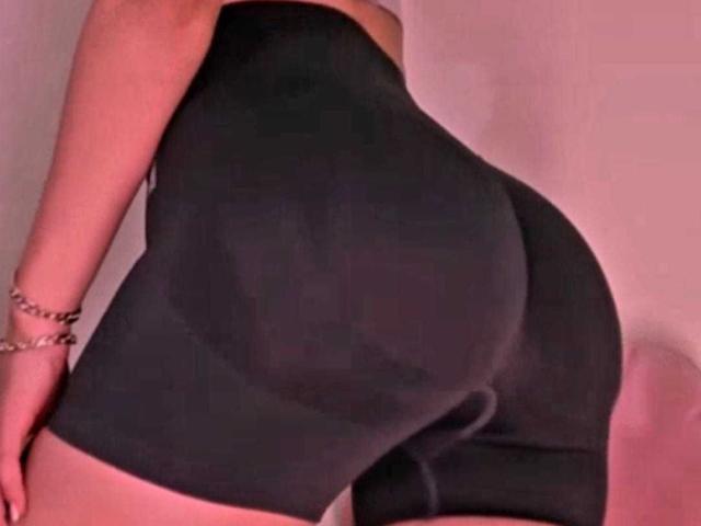 Close-up of BlueNightX's lower body. She wears tight, black, form-fitting shorts that highlight her large, round buttocks. Her right arm, adorned with a silver bracelet, is slightly bent. The image has a pinkish, warm tone. The texture of the shorts is smooth, contrasting with the soft, slightly dimpled skin of her buttocks. The angle emphasizes her curves, with the shorts clinging to her shape.