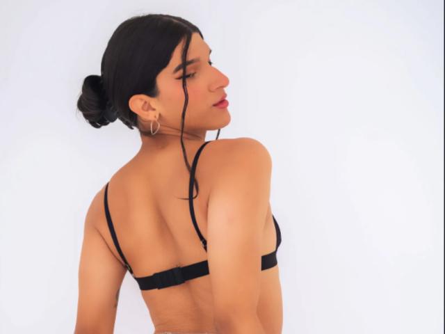 BiancaBaldwin with black bra, butt facing camera, arms down, dark hair in bun, red lipstick, small hoop earrings, white background, soft light, slight smile.
