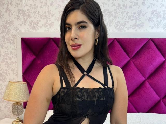 JasminSwit with dark hair, wearing a black lace and strapless top, sitting on a bed with a purple, diamond-patterned headboard, and a gold lamp on the left.