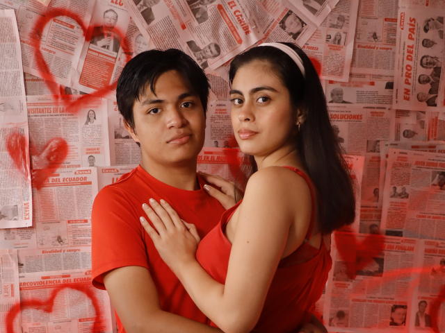SofiNdLogan with black hair, red shirt, and medium build, holds a curvy woman with brown hair, white headband, red dress, and large tits, against a newspaper wall with red hearts.