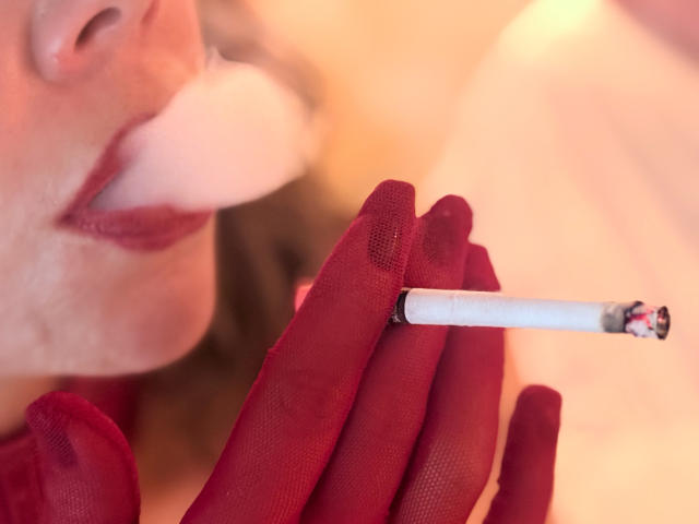 EmiliaMilf's red-gloved hand holds a cigarette, blowing a thick white smoke into her pouty red-lipsticked mouth. Her face is partially visible, showing soft skin and light brown hair.