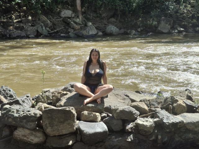 KatherinLatin with tits in a dark tank top, sitting cross-legged on rocky riverbank, brown water flowing behind, forest in background, sunlight highlighting her long brown hair and tan skin.