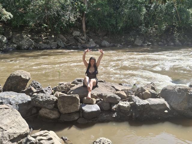 KatherinLatin, with small tits and a black bikini, sits on rocky riverbed, arms raised, in sunny forest. Water flows around her, green trees in background.