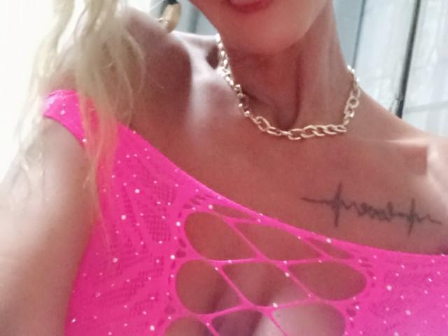 Meddusa69 with blonde hair, wearing a sparkling pink top with cutouts, revealing small tits, and a gold chain necklace, has a heartbeat tattoo on her right chest.