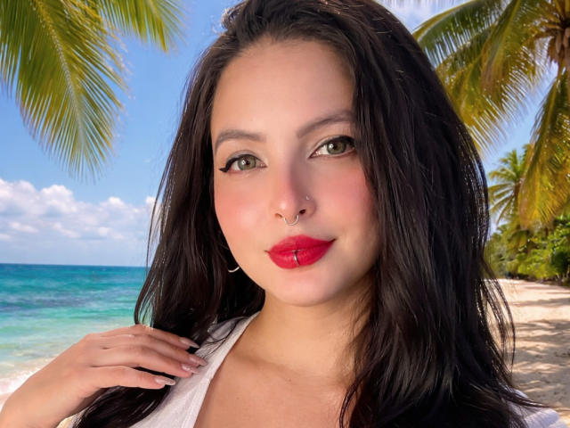 SofiaLeal with dark hair, green eyes, red lipstick, nose ring, and white top, standing on a beach with palm trees and clear water in the background.