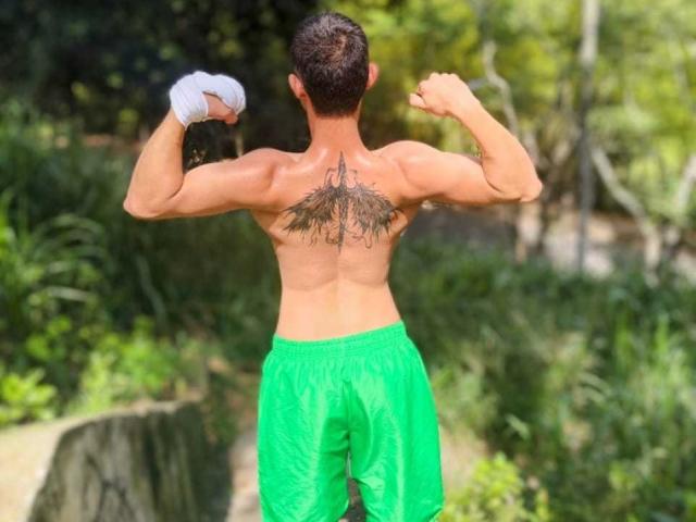 DynamicCoulee's muscular back with a large, intricate tattoo between the shoulders, flexing both arms, wearing green shorts, and a white wristband in a sunny, green forest.