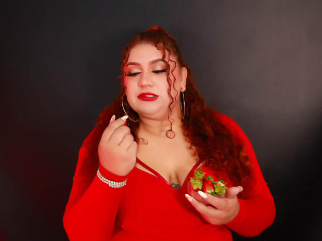 SeleneFhox69 with curly red hair, red lipstick, red top, holding strawberries, wearing hoop earrings, and a diamond bracelet, looking down with a slight smile, against a dark background.