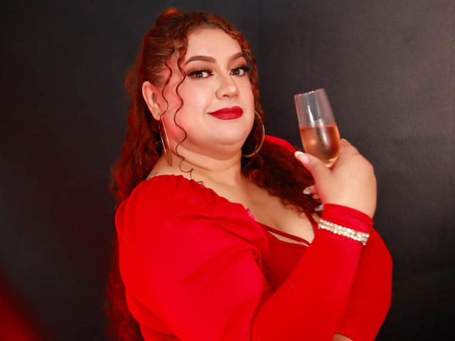 SeleneFhox69 with curly red hair, red lipstick, wearing a red dress, holding a glass of champagne, large hoop earrings, and a pearl bracelet, smiling confidently.
