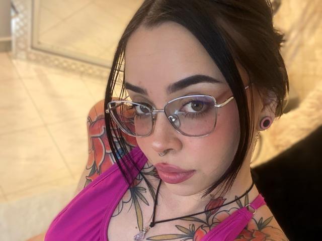 NaiaWhisper with glasses, nose ring, and pink lipstick, showing colorful tattoos on her tits and neck, wearing a pink top, and black ear gauges, in a selfie.