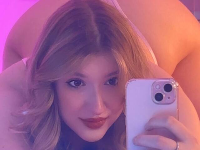A selfie of Synrix with wavy blonde hair and light skin, looking at the camera with a slight smile. She's holding a white iPhone with a silver frame in her right hand. Her eyes are dark and she's wearing red lipstick. The background is bathed in purple and orange light, creating a soft, warm glow. Her left arm is partially visible, and she's leaning forward. The image has a casual, intimate feel.