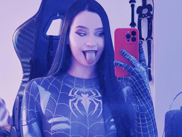 MaryLori takes a selfie in a mirror. She has long, straight black hair and a fair skin tone. She's wearing a Spider-Man costume with a large spider emblem on the chest, and black, web-patterned gloves. Her tongue is out, and she's looking at her red phone with three cameras. The chair behind her is black with a cutout in the back. The image has a blue tint.