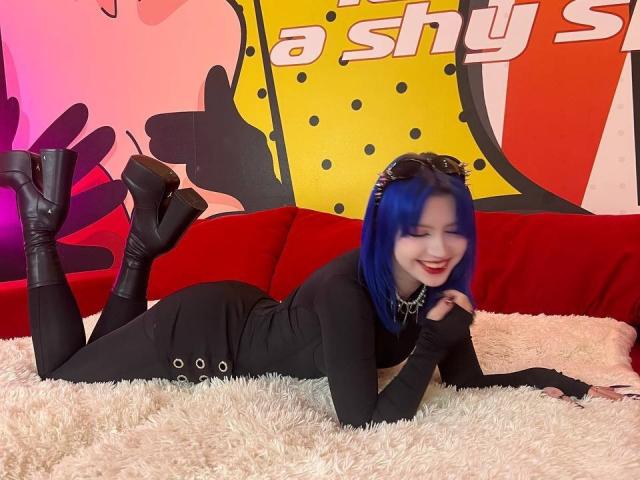 SelenaIris with blue hair, black long-sleeve dress, black boots, and chain necklace, lying on a fluffy white rug, smiling, showing her ass. She's on a red couch.