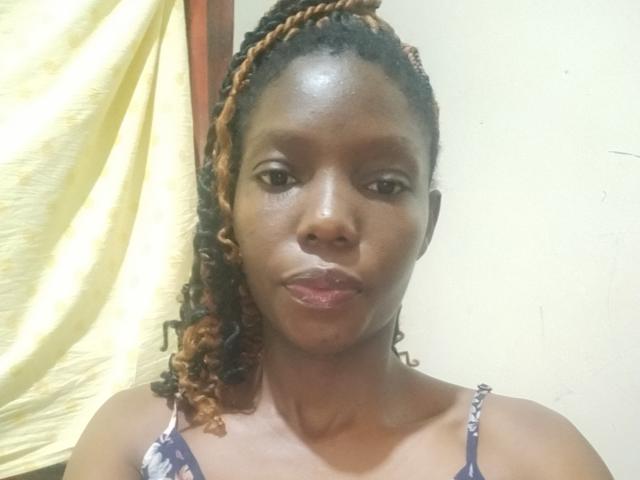 JulianaMirabel has dark brown skin, curly brown hair with blonde tips, and is wearing a purple floral top, with a neutral expression and slightly shiny face.