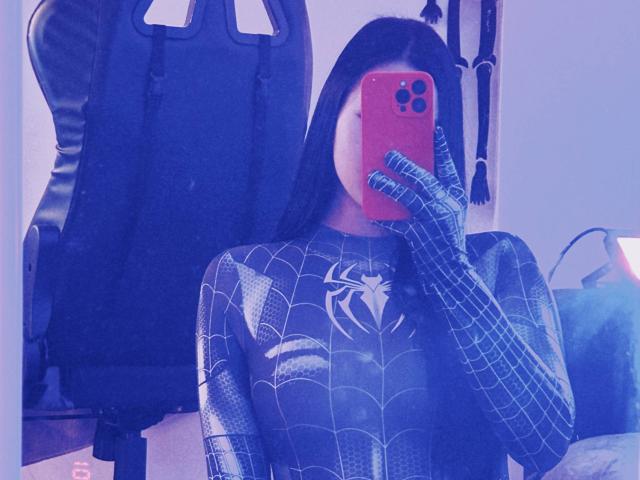 MaryLori in Spider-Man cosplay, blue-tinted mirror selfie, black web-patterned suit, gloves, red phone, black chair, long straight black hair, holding phone covering face.