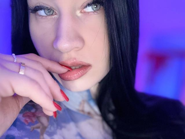 MaryLori with pale skin, black hair, glossy pink lips, and red nails, touching her lip with a finger that has a silver ring. Her eyes are blue and she's looking up.