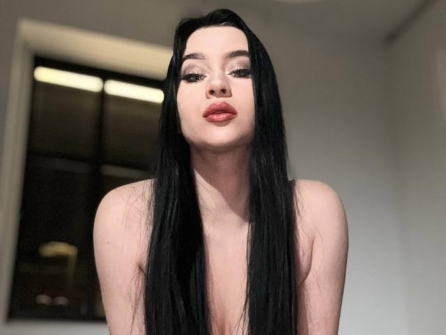MaryLori has long black hair, pale skin, and red lipstick. She's topless with her tits visible, looking down with a slight pout. Her eyelashes are long and dark.